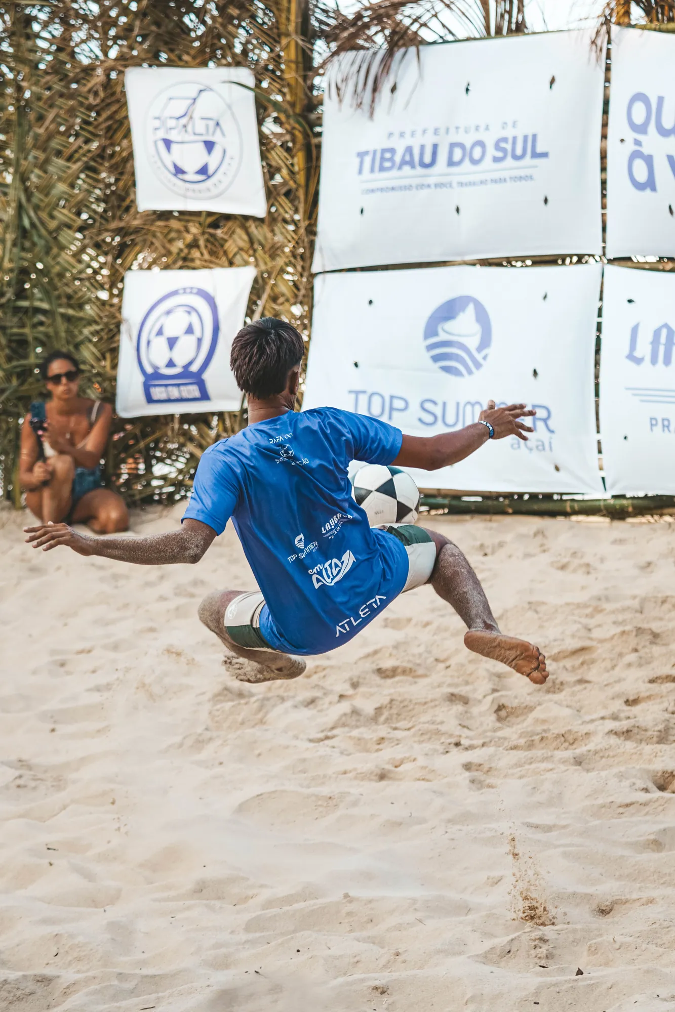 Jogador de camisa azul dando um voleio na areia